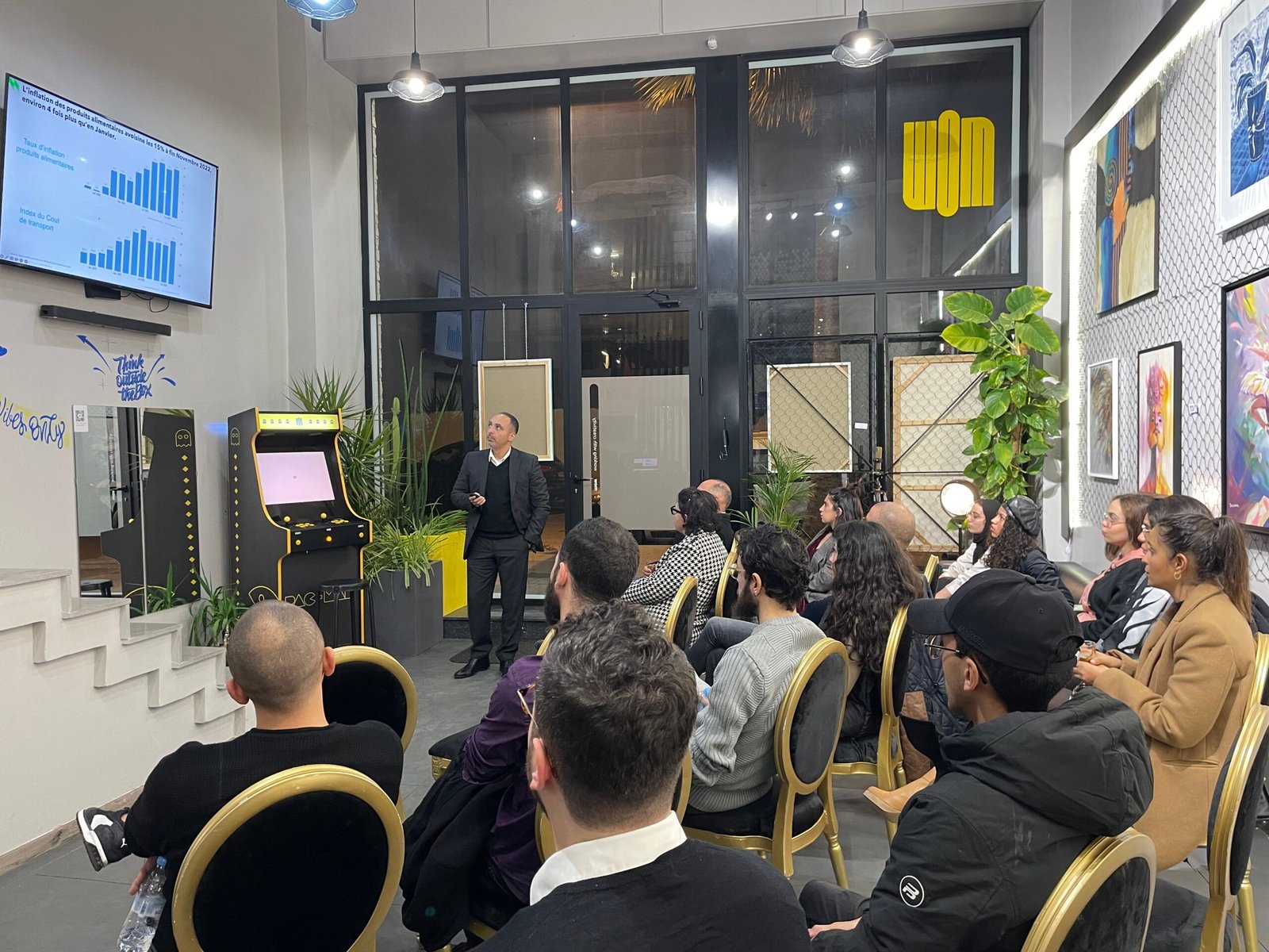 Présentation en direct par un leader au sein de Mow devant un auditoire attentif, avec des graphiques de données projetés, dans un espace de travail dynamique et artistiquement décoré