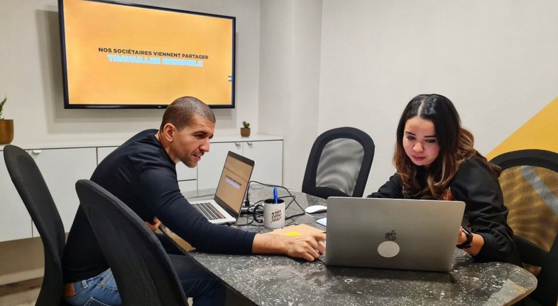 Deux partenaires d'affaires travaillant sur des ordinateurs portables dans une salle de conférence chez Mow avec un écran affichant 'Nos sociétaires viennent partager